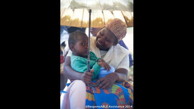 A Mother shelters from the sun under an umbrella and smiles at her disabled child who is sitting on her lap