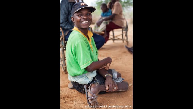 Another physically disabled man smiles at the camera while he sits on the floor