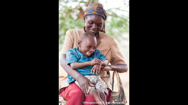 A young boy who has a physical disability laughs with his Mother as he sits on her lap