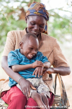 A young boy who has a physical disability laughs with his Mother as he sits on her lap