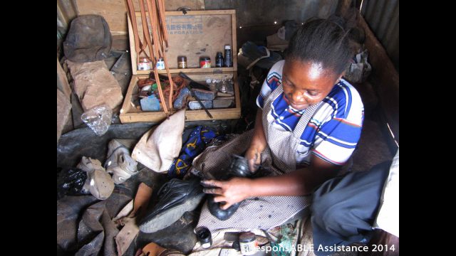 Margaret sits on the floor of her roadside shack surrounded by the tools and materials she uses to mend and clean people’s shoes
