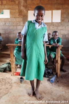 A young girl with a physical disability stands shyly on the dirt floor of her classroom